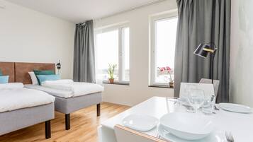 Une chambre d’hôtel avec deux lits, une table à manger dressée avec des assiettes et des verres, et une vue sur l’extérieur grâce à de grandes fenêtres.