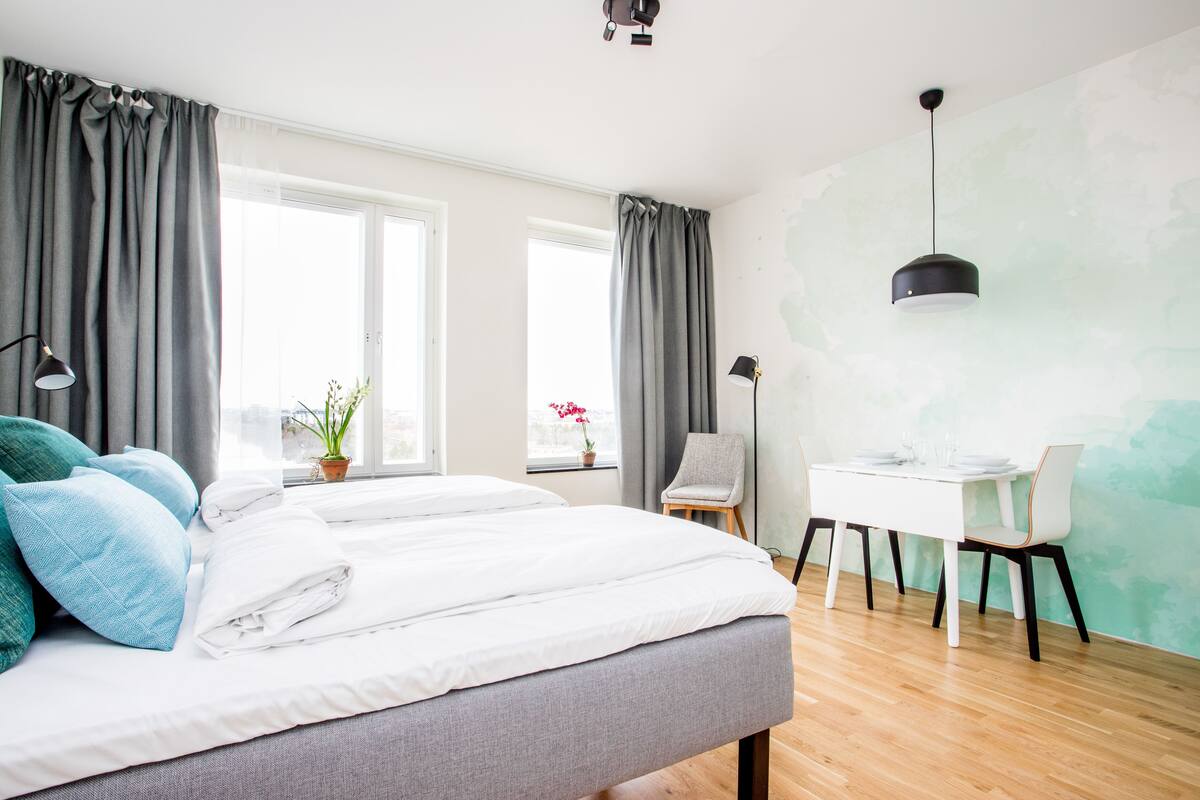 Un dormitorio moderno con una cama grande, dos ventanas con cortinas, un escritorio con sillas y una lámpara colgante.