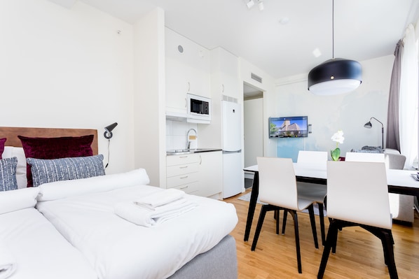 A modern hotel room with a bed, a desk, a dining table, and a kitchen area.