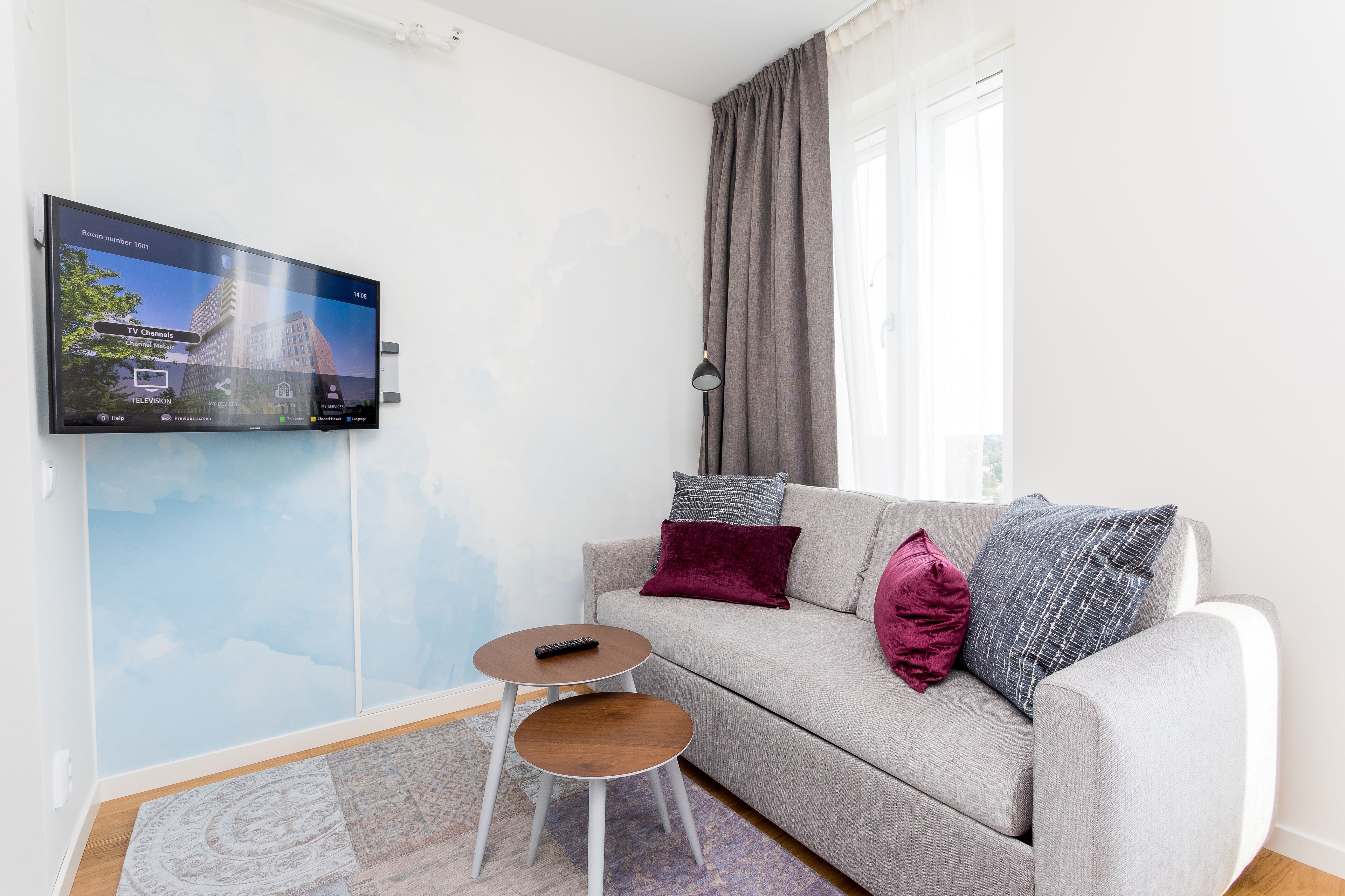 Ett modernt vardagsrum med en grå soffa, två sidobord, en väggmonterad TV och en mönstrad matta.