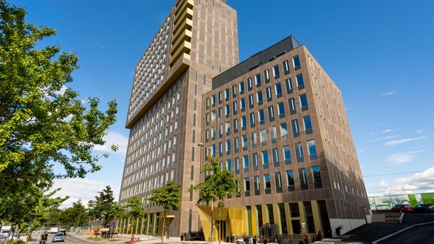 A modern high-rise building with multiple floors, large windows, and a prominent corner entrance. - ApartDirect Älvsjö (Älvsjö)