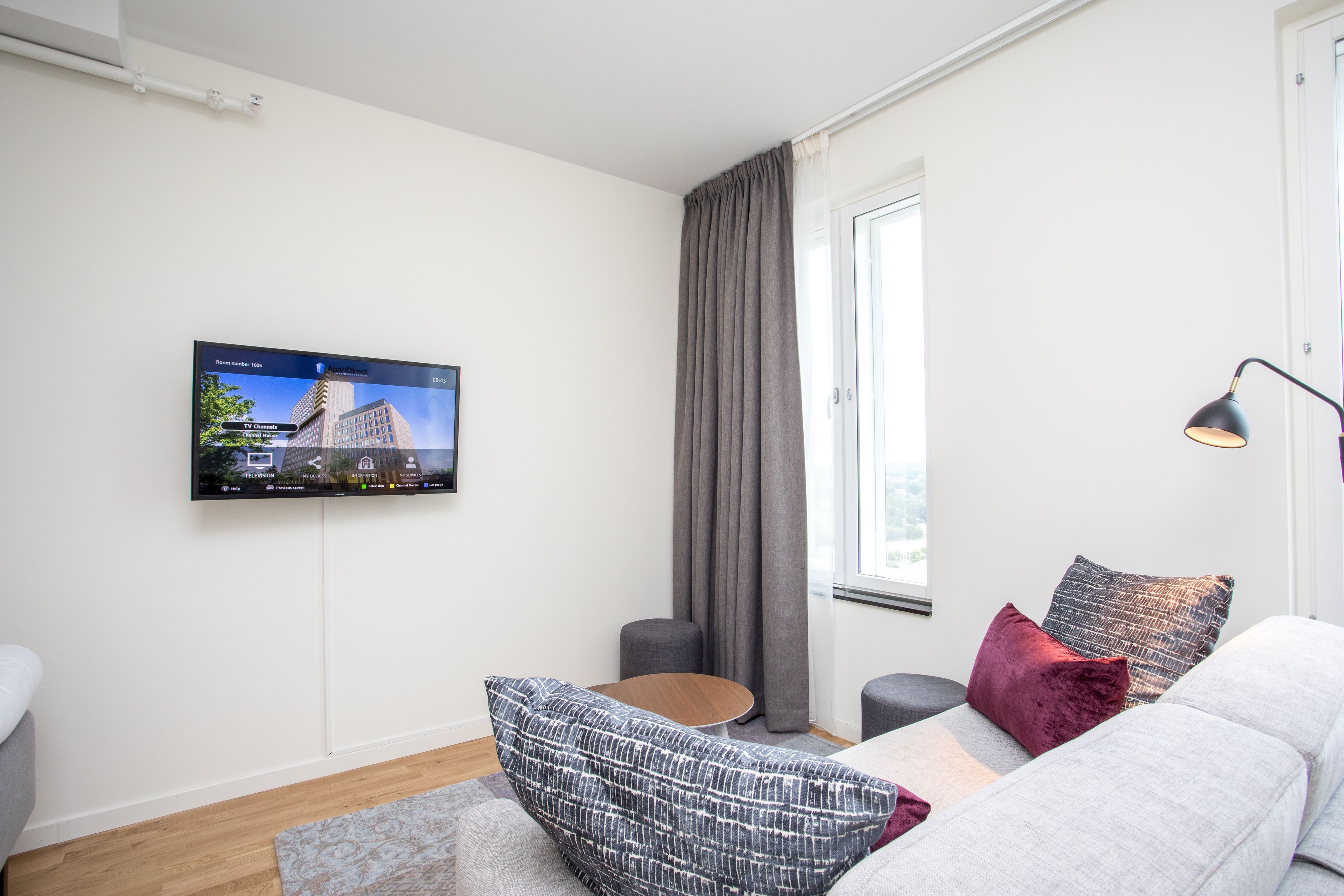 A modern living room with a flat-screen TV mounted on the wall, a grey sofa with patterned cushions, a round coffee table, and a window with grey curtains.