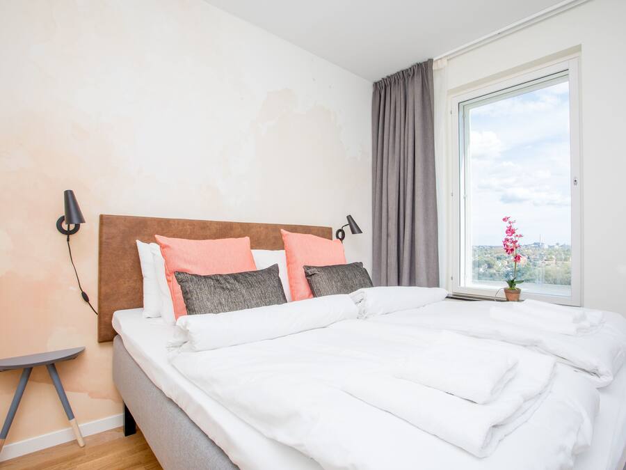 A neatly made bed with white linens, a headboard with a brown panel, and a window with a view of a cityscape.