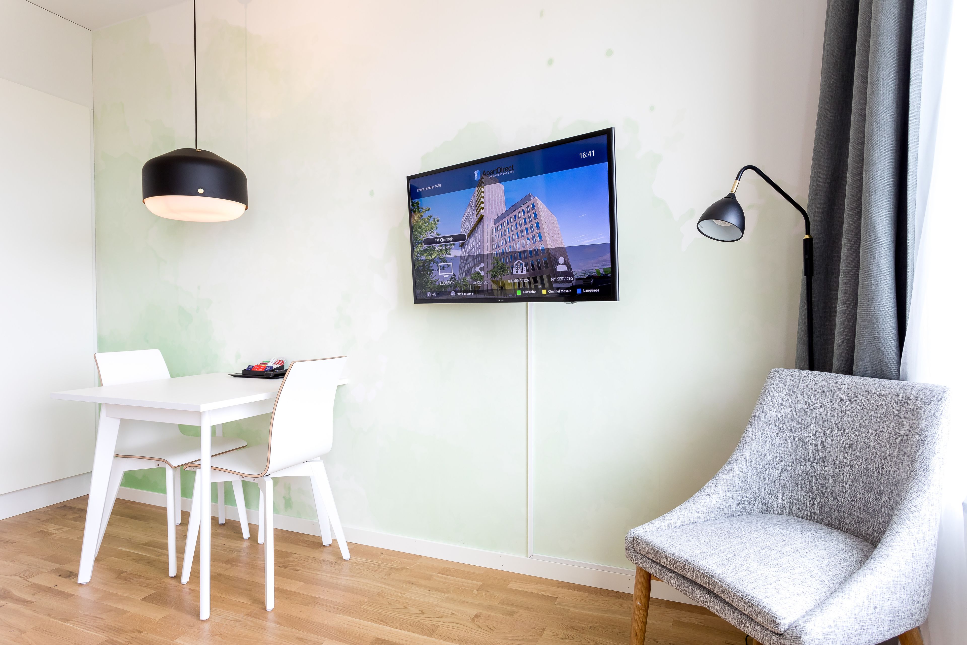A modern room with a flat-screen TV mounted on the wall, a white table with chairs, a grey armchair, and a black pendant light.