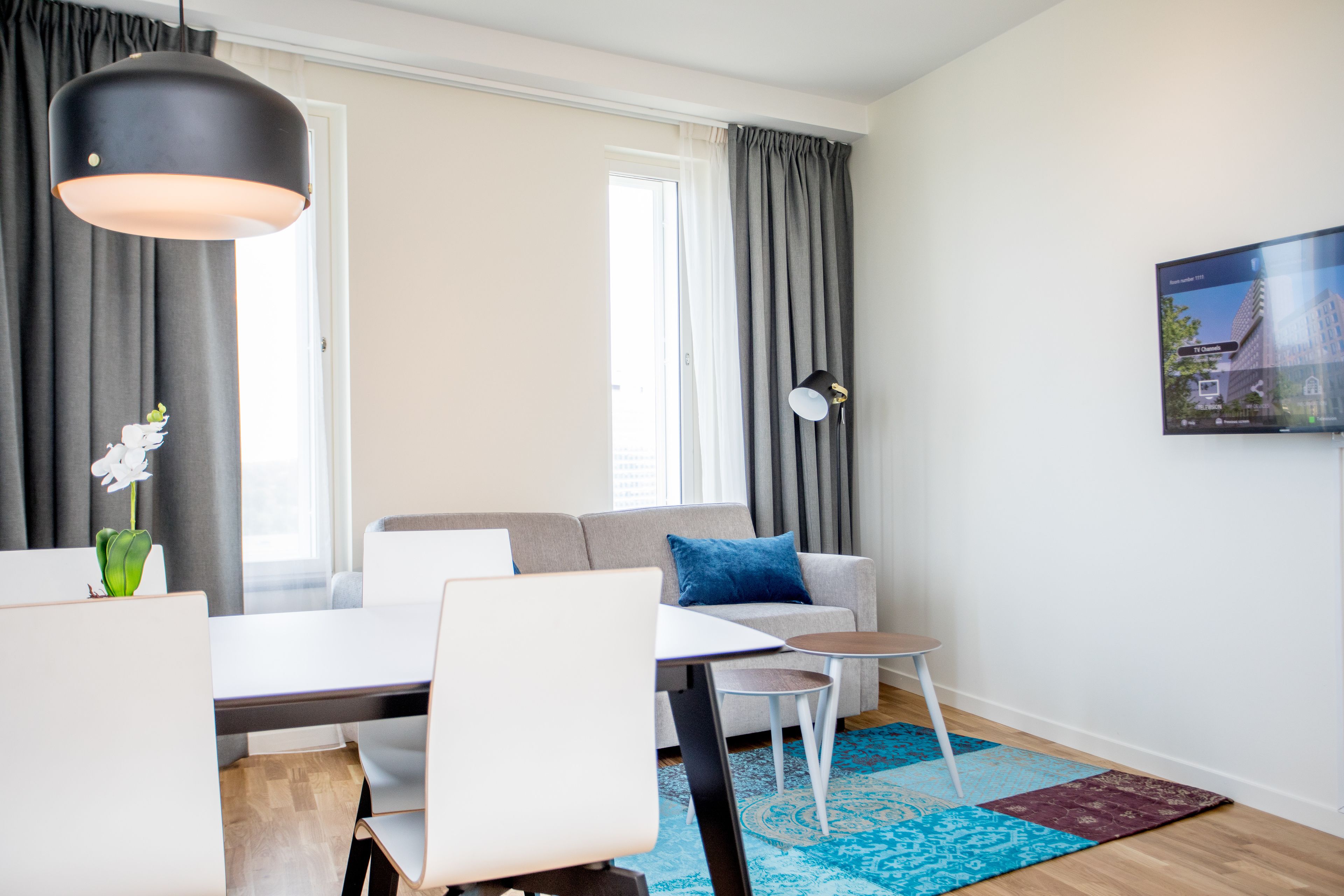 Een moderne woonkamer met een bank, een eettafel en stoelen. Er is een groot raam met gordijnen, een aan de muur bevestigde televisie en een decoratieve plant.