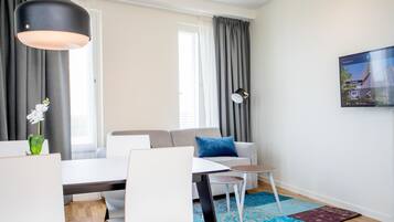Een moderne woonkamer met een bank, een eettafel en stoelen. Er is een groot raam met gordijnen, een aan de muur bevestigde televisie en een decoratieve plant.