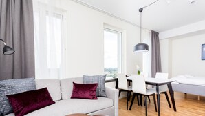 A modern living room with a sofa, two round tables, a dining area with chairs, and a large window providing a view. - ApartDirect Älvsjö (Älvsjö)