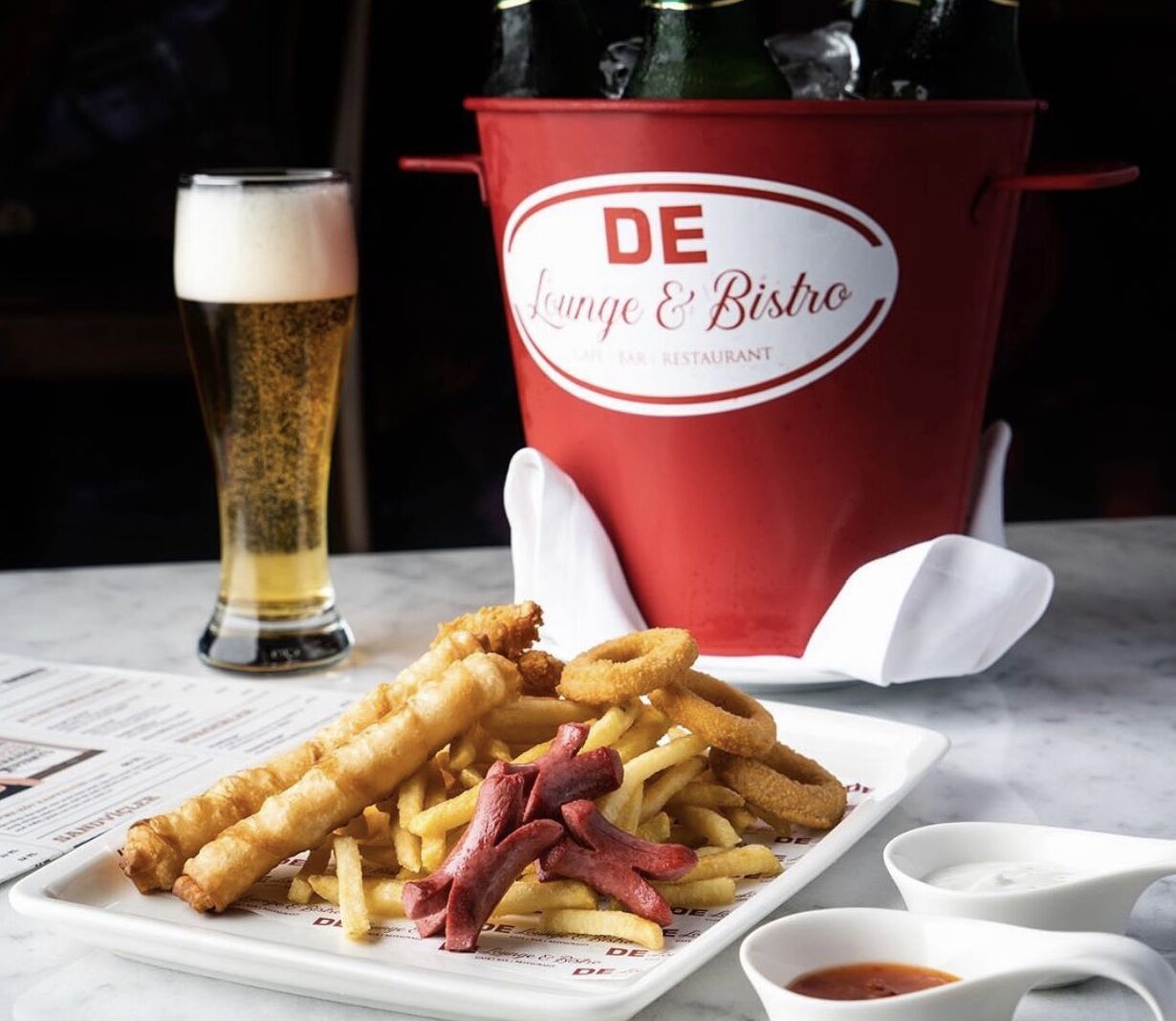 A plate of food with fries and fish and chips, a glass of beer, and a red bucket labeled "DE Lounge & Bistro".