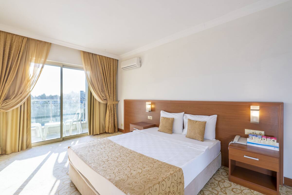 Habitación de hotel con una cama grande, una mesita de noche con teléfono y artículos de aseo, y vistas de la ciudad a través de grandes ventanales.