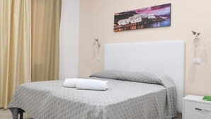 Ein ordentlich bezogenes Bett mit einer grau gemusterten Tagesdecke und zwei Kissen, ein weißer Nachttisch mit Schublade sowie ein an der Wand hängendes Stadtbild.