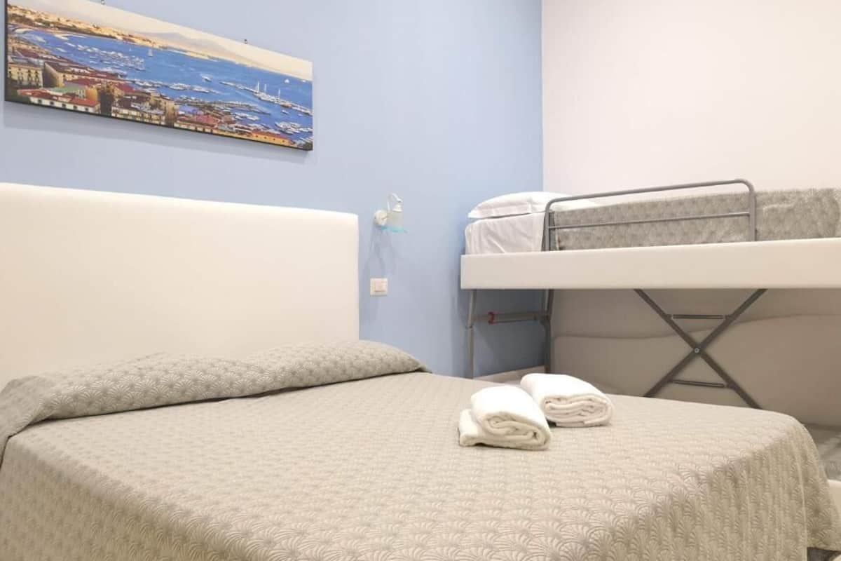 Una habitación de hotel con cama, literas, un cuadro de paisaje costero y una mesita de noche con lámpara.
