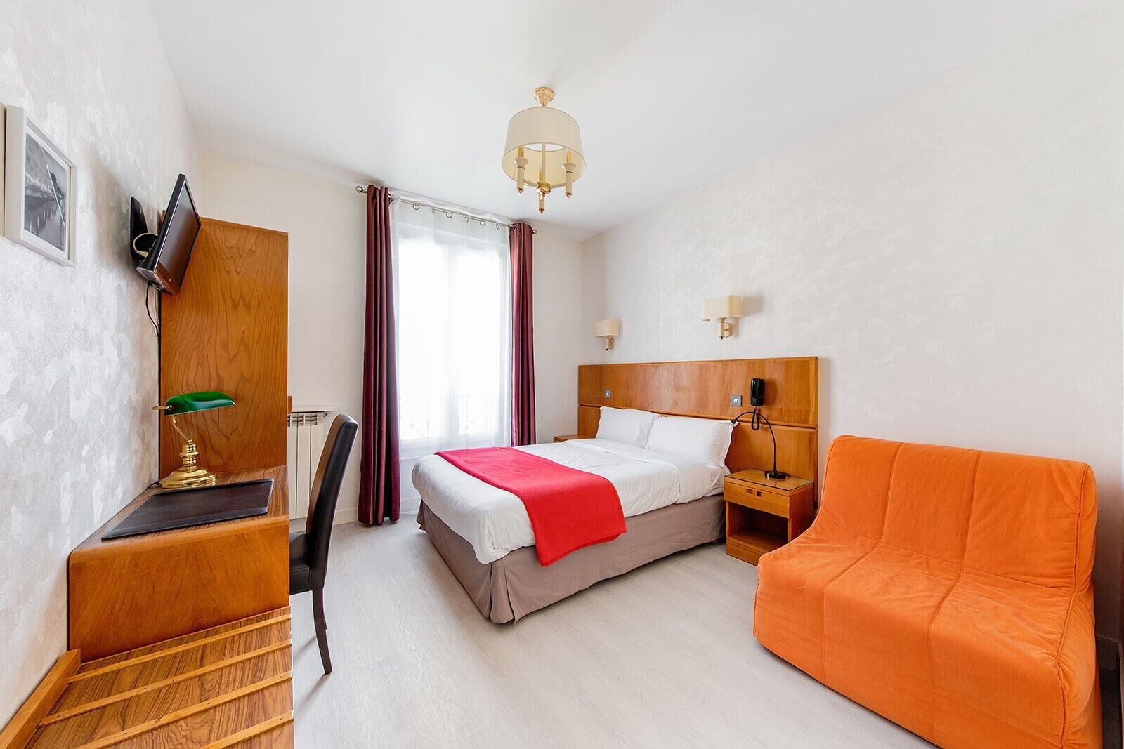 A hotel room with a bed, a desk with a lamp, a chair, a nightstand with a phone, and an orange couch.