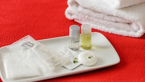 A white tray with toiletries on a red towel, a folded white towel, and a bed with white bedding. - Crystal Hotel (Levallois-Perret)