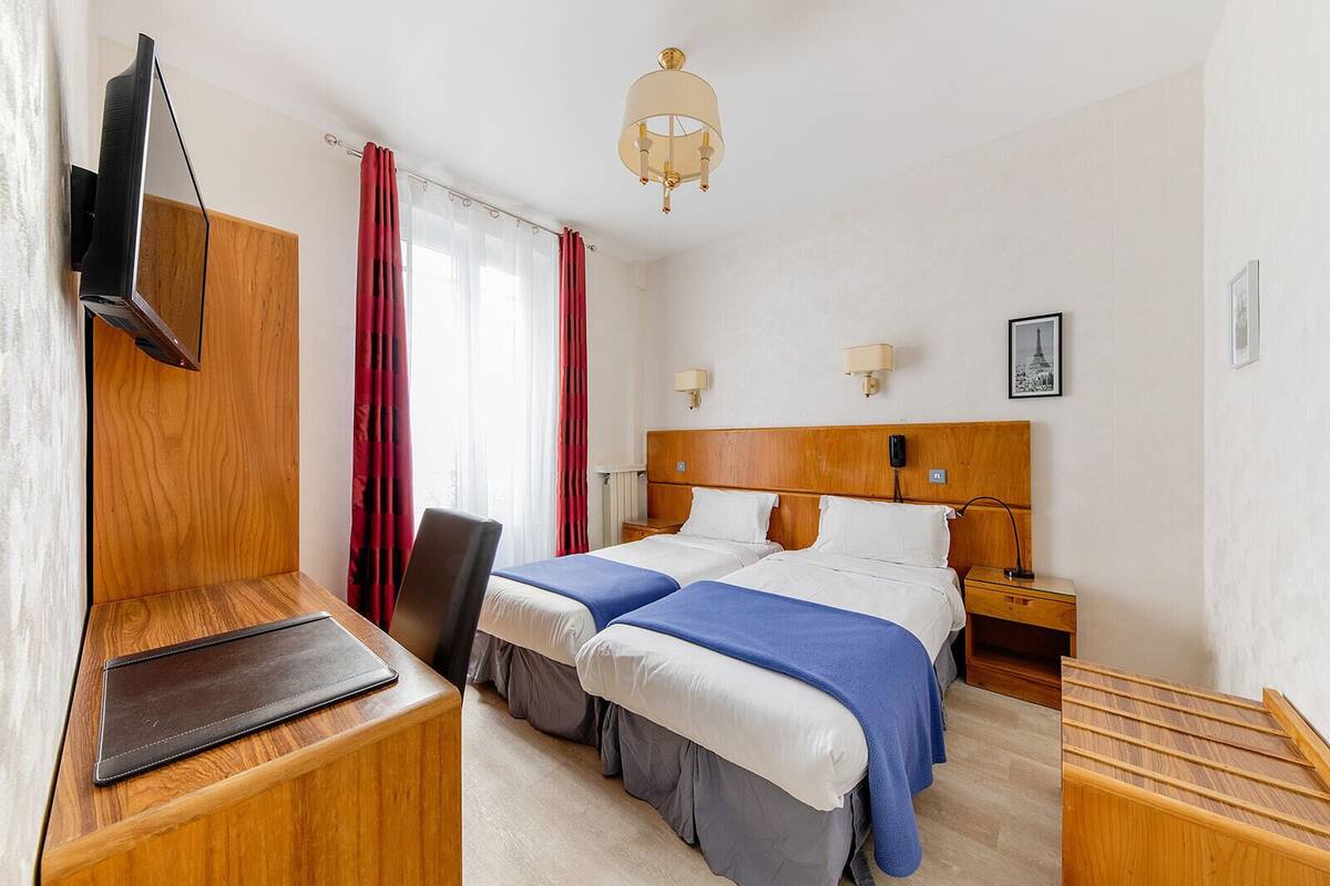 A hotel room with a wooden headboard, two beds with blue and white bedding, a desk with a laptop, a TV mounted on the wall, a bedside lamp, and a picture of the Eiffel Tower on the wall.