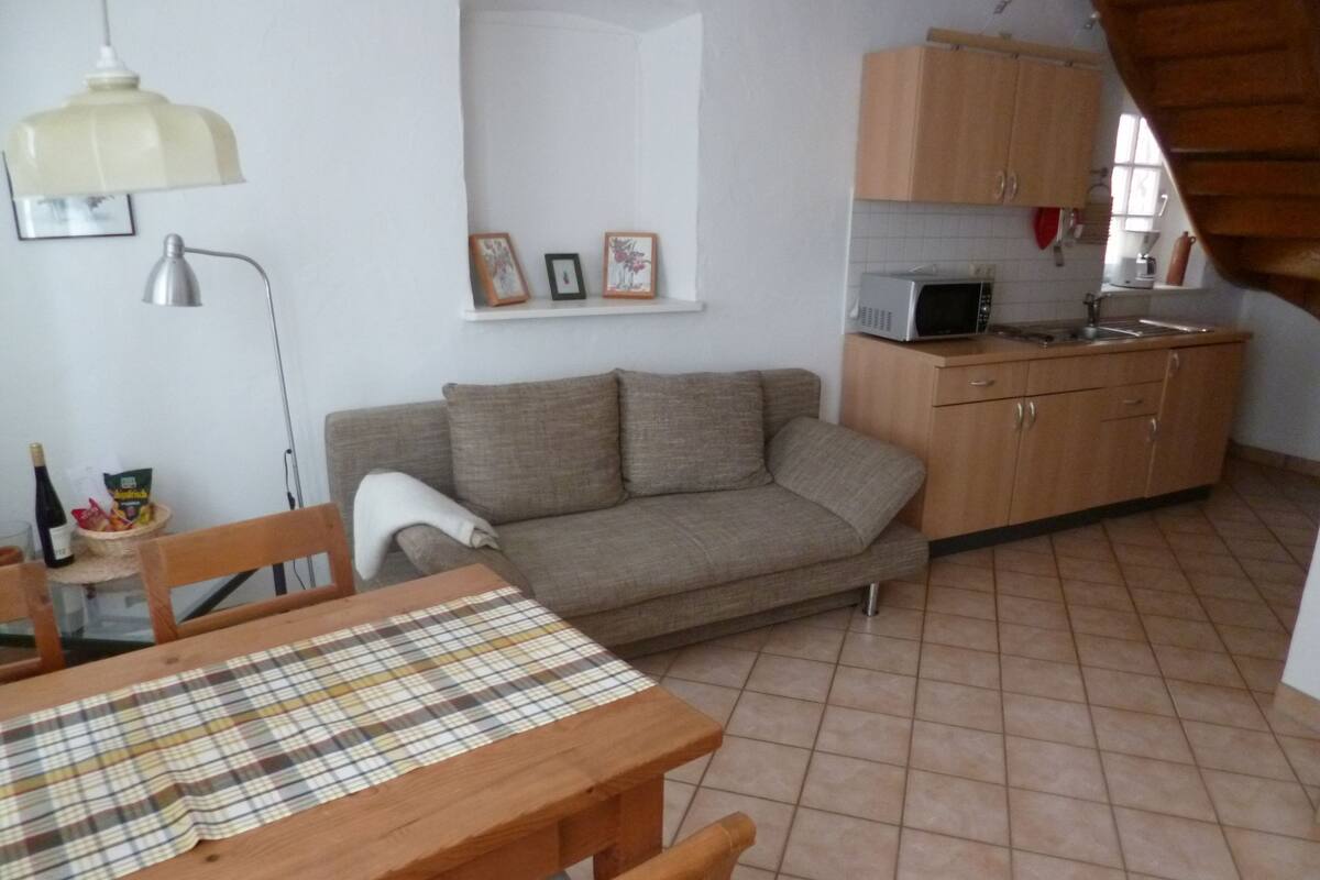 Un espace de vie compact comprenant un canapé, une table à manger recouverte d’une nappe à carreaux, un meuble en bois et une petite cuisine équipée d’un four à micro-ondes.