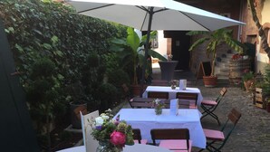 A patio area with round tables and chairs, covered by umbrellas, and surrounded by greenery and potted plants. - Alter Winzerhof Weisenheim am Berg e.K. (Weisenheim am Berg)