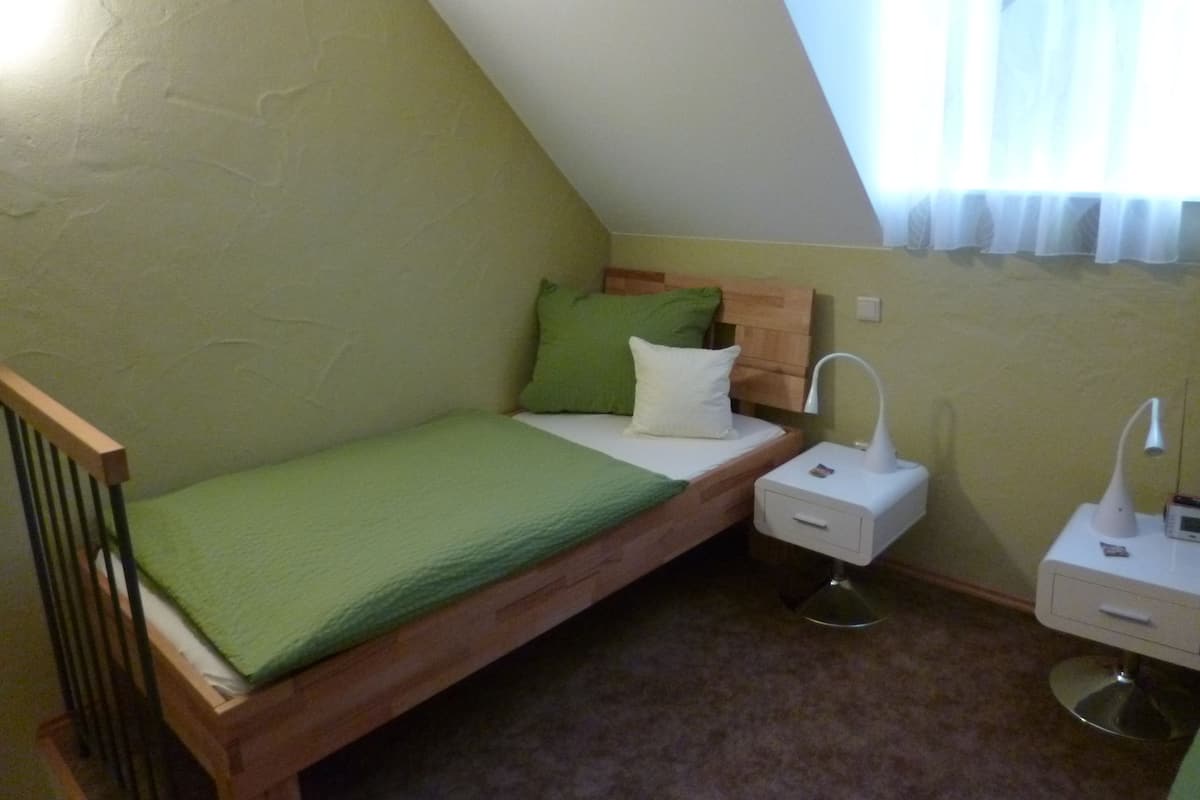 Une petite chambre avec un lit simple, un plafond en pente, une tête de lit en bois, un couvre-lit vert, une lampe de chevet, une table de chevet et une petite commode.