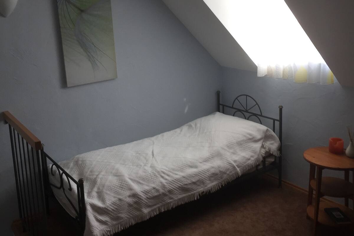 Une petite pièce faiblement éclairée, avec un lit simple, une petite table et un plafond en pente percé d’une lucarne.
