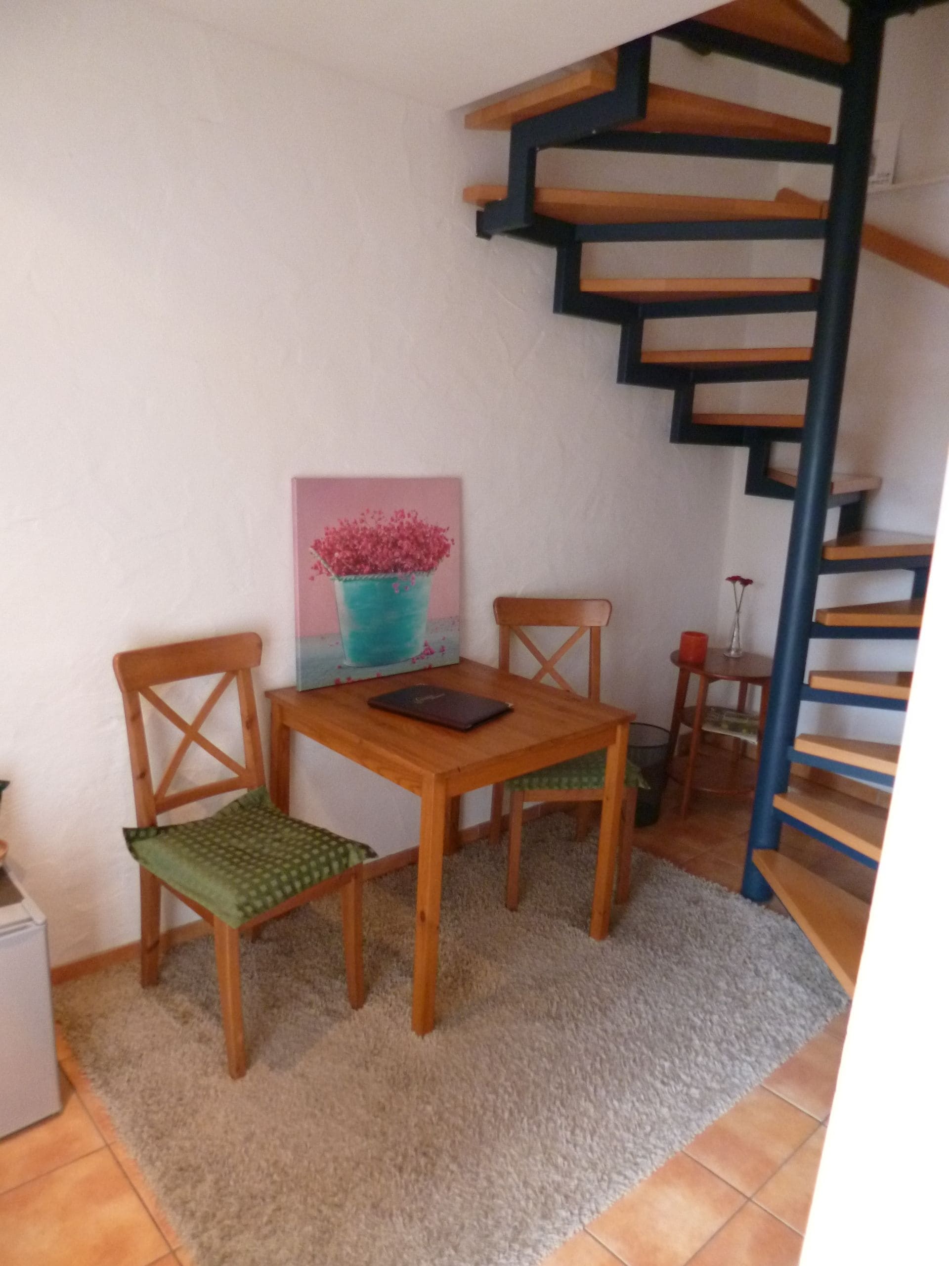 Un petit coin repas avec une table et des chaises en bois, un tableau au mur et un escalier en colimaçon.