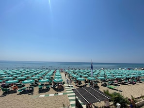 A beach with numerous green umbrellas and lounge chairs, a clear sky, and the ocean in the background.