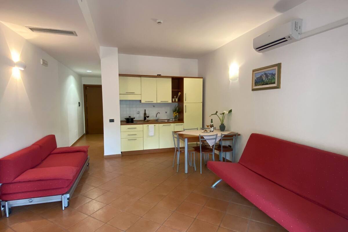 A living room with a red sofa, a dining table with chairs, and a kitchen area in the background.
