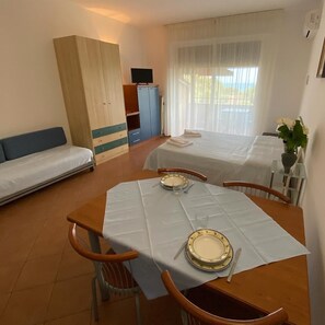 Ein Hotelzimmer mit zwei Betten, einem Esstisch für zwei Personen, einem Fernseher und einem Fenster mit Vorhängen.