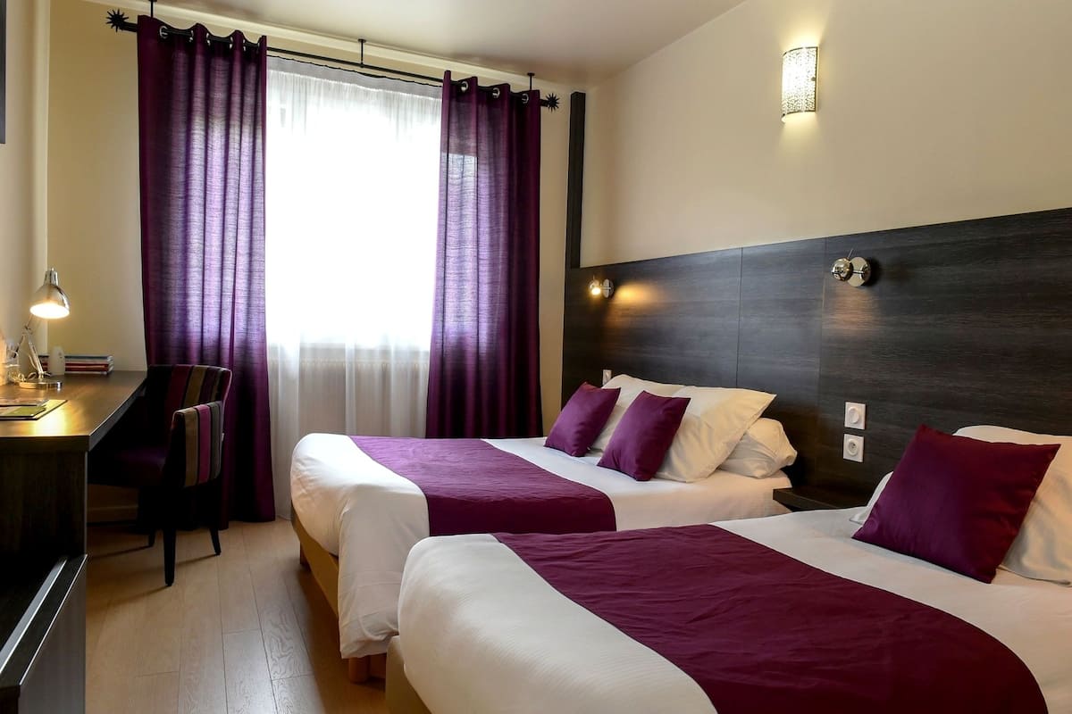 A hotel room with two beds, a desk, a chair, and a window with purple curtains.