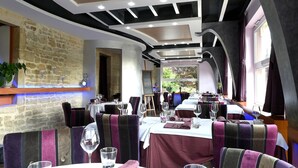 Een restaurant met een stenen muur, een houten plafond en gestreept beklede zitbanken. De tafels zijn gedekt met wijnglazen en bestek.