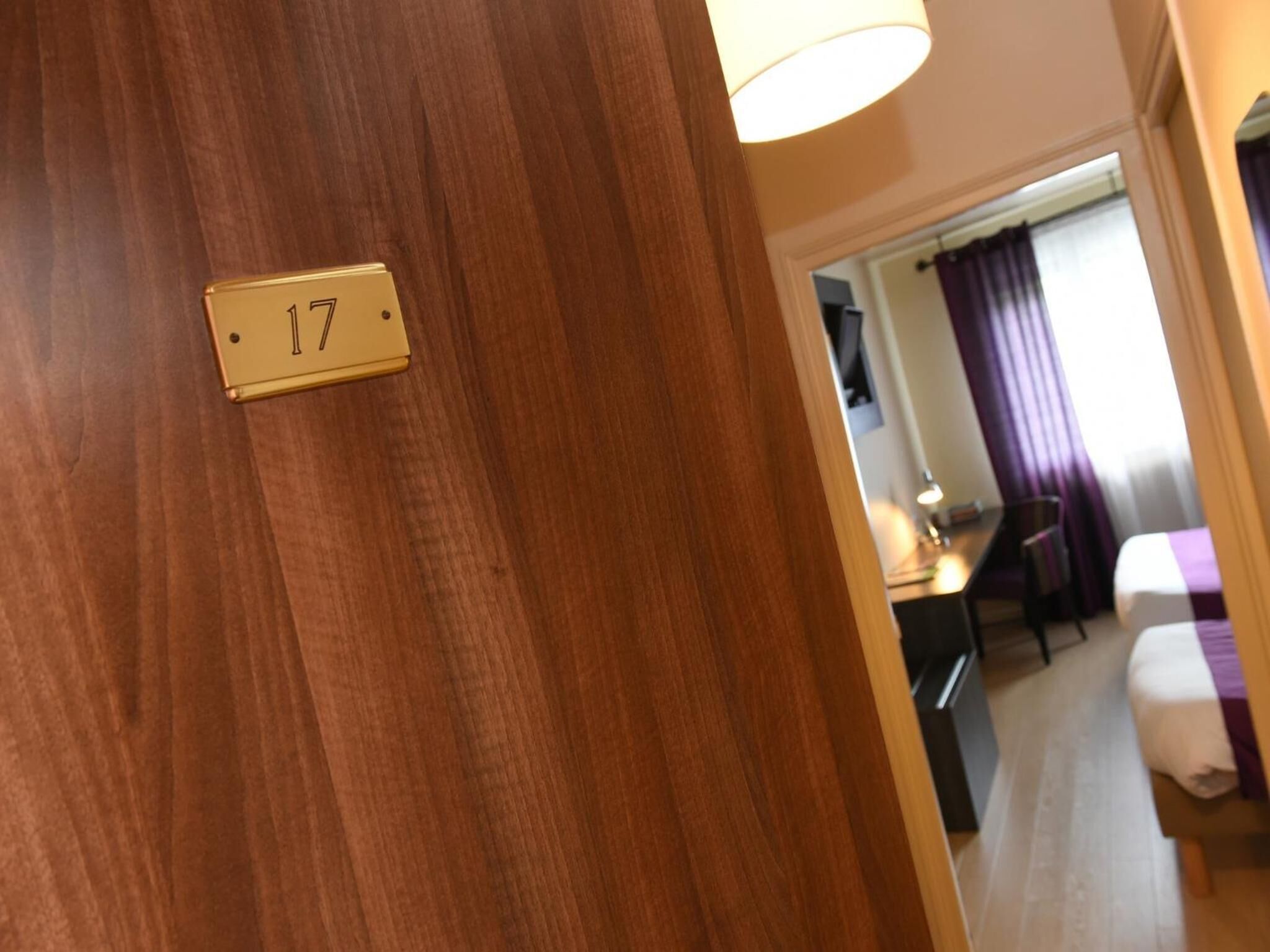 A hotel room with a wooden door, a number plate indicating room 17, a bed with a purple blanket, a desk with a lamp, and a window with purple curtains.