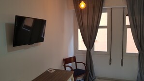 A room with a flat-screen TV mounted on the wall, a wooden desk with a vase of flowers, a chair, and a window with curtains. - Fan Rooms Duquesa (Málaga)