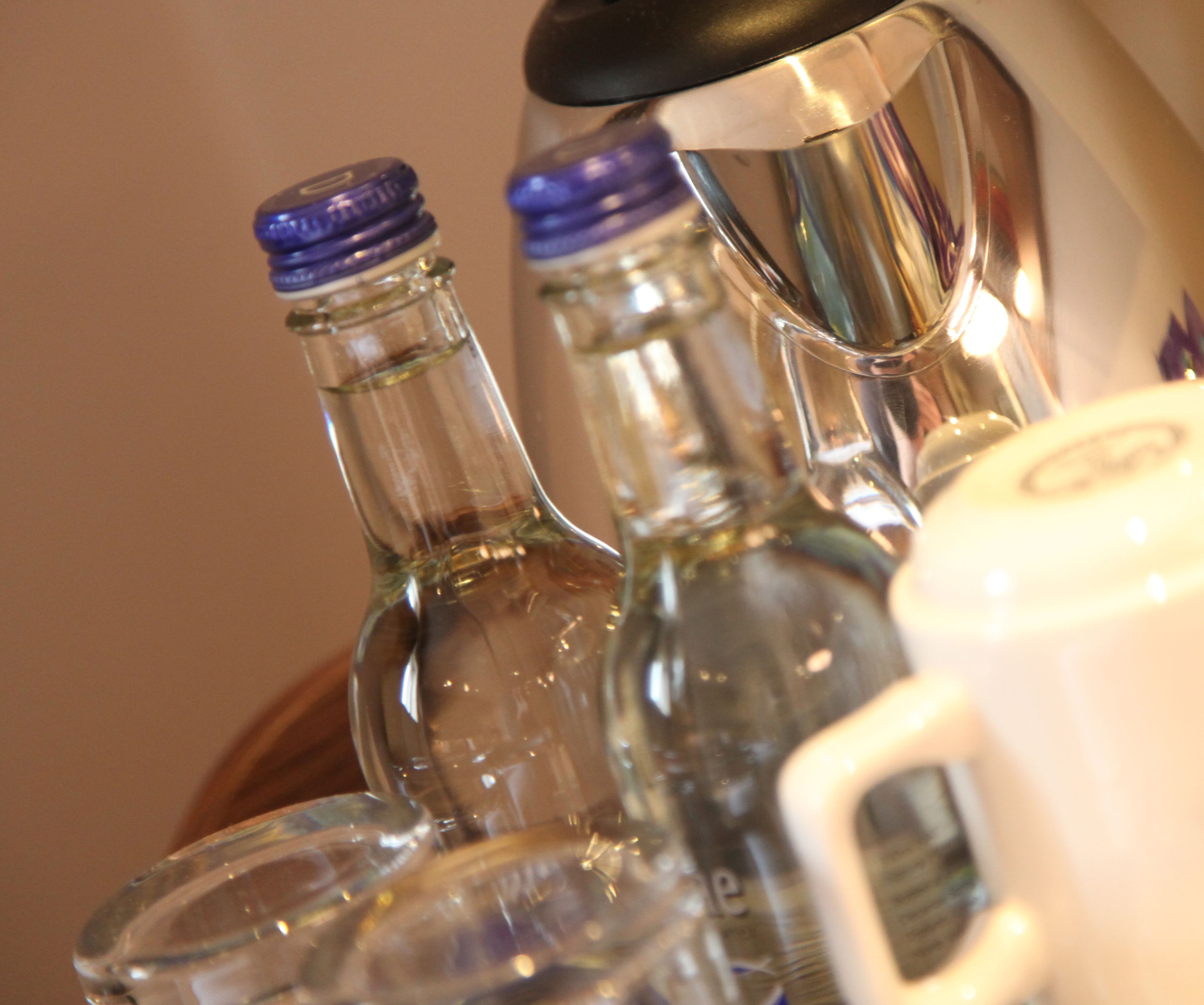 Primer plano de varias botellas de vidrio con tapones azules, probablemente conteniendo bebidas, y un recipiente blanco con un logo, posiblemente para servir o guardar.