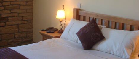 A neatly made bed with white linens and a brown throw blanket, a wooden headboard, a nightstand with a lamp, and a stone wall.