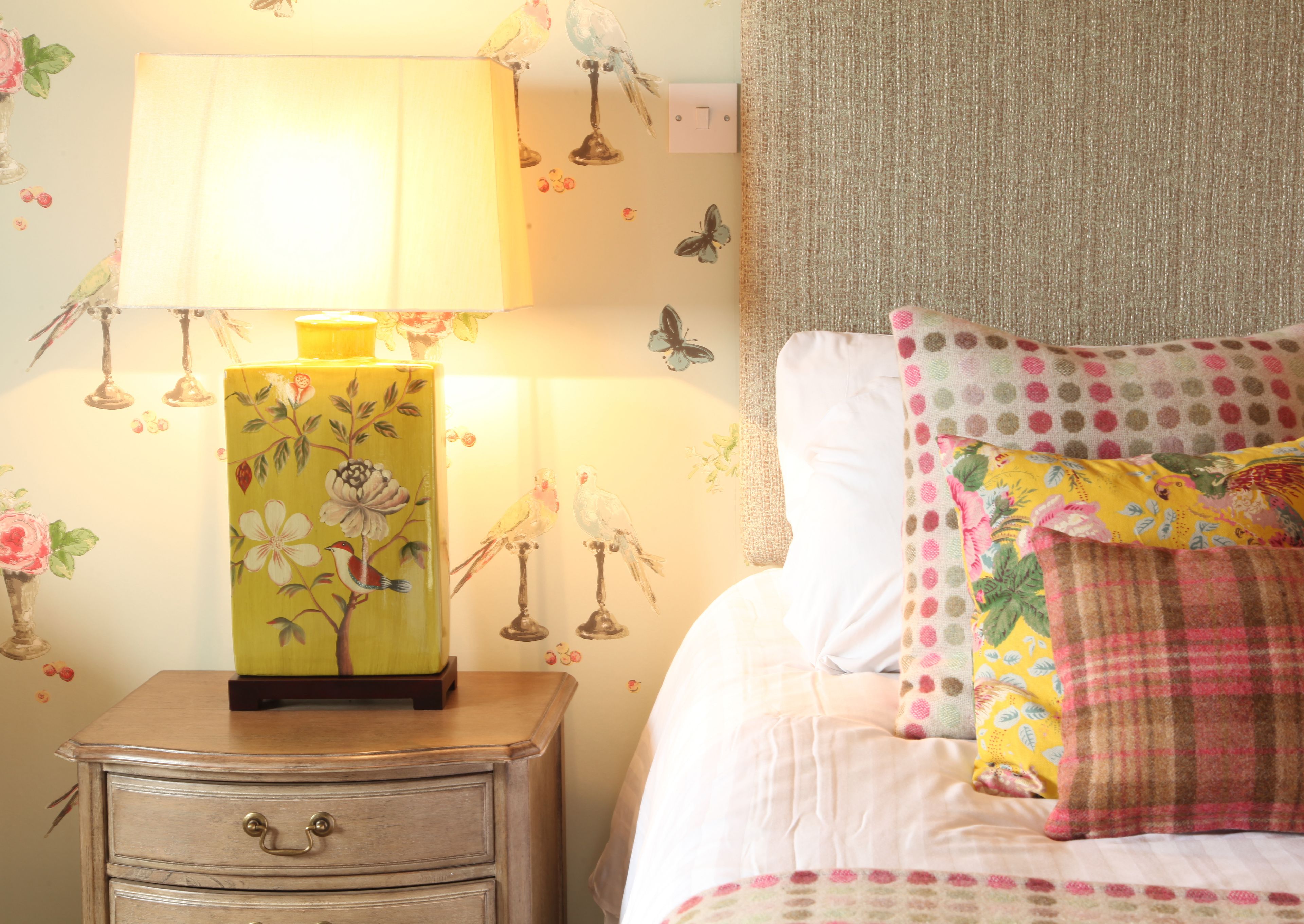 A bedroom with a nightstand, a lamp, a bed with a floral pillow, and wall decorations including birds and butterflies.