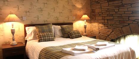 A neatly made bed with a checkered blanket, two pillows, and folded towels. There are two bedside lamps on wooden nightstands. The room features a stone wall and wallpaper with a repeating pattern.