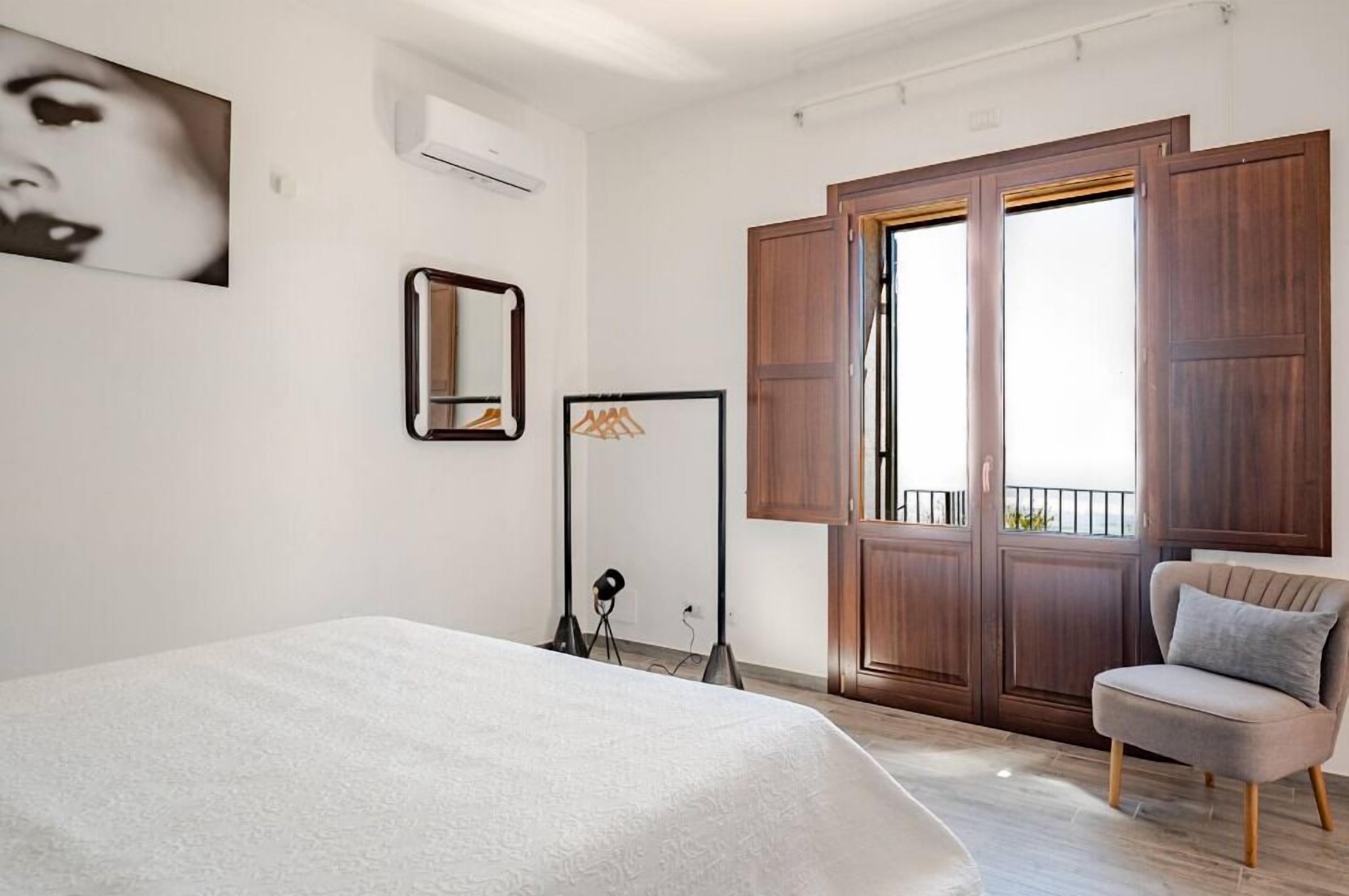Ein Schlafzimmer mit einem Bett, einem Sessel, einem Kleiderschrank, einem Spiegel und einem Fenster.