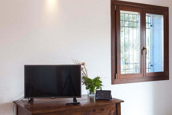 A television on a wooden stand, a remote control, and a small potted plant on a white wall. - Etnella enoagriturismo Notti Stellate (Mascali)