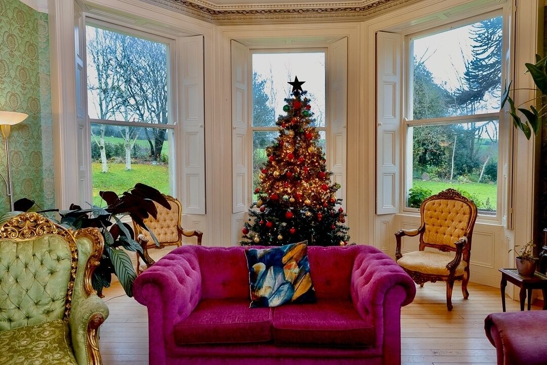 A room with a large window, a Christmas tree, a red sofa, and two antique chairs.