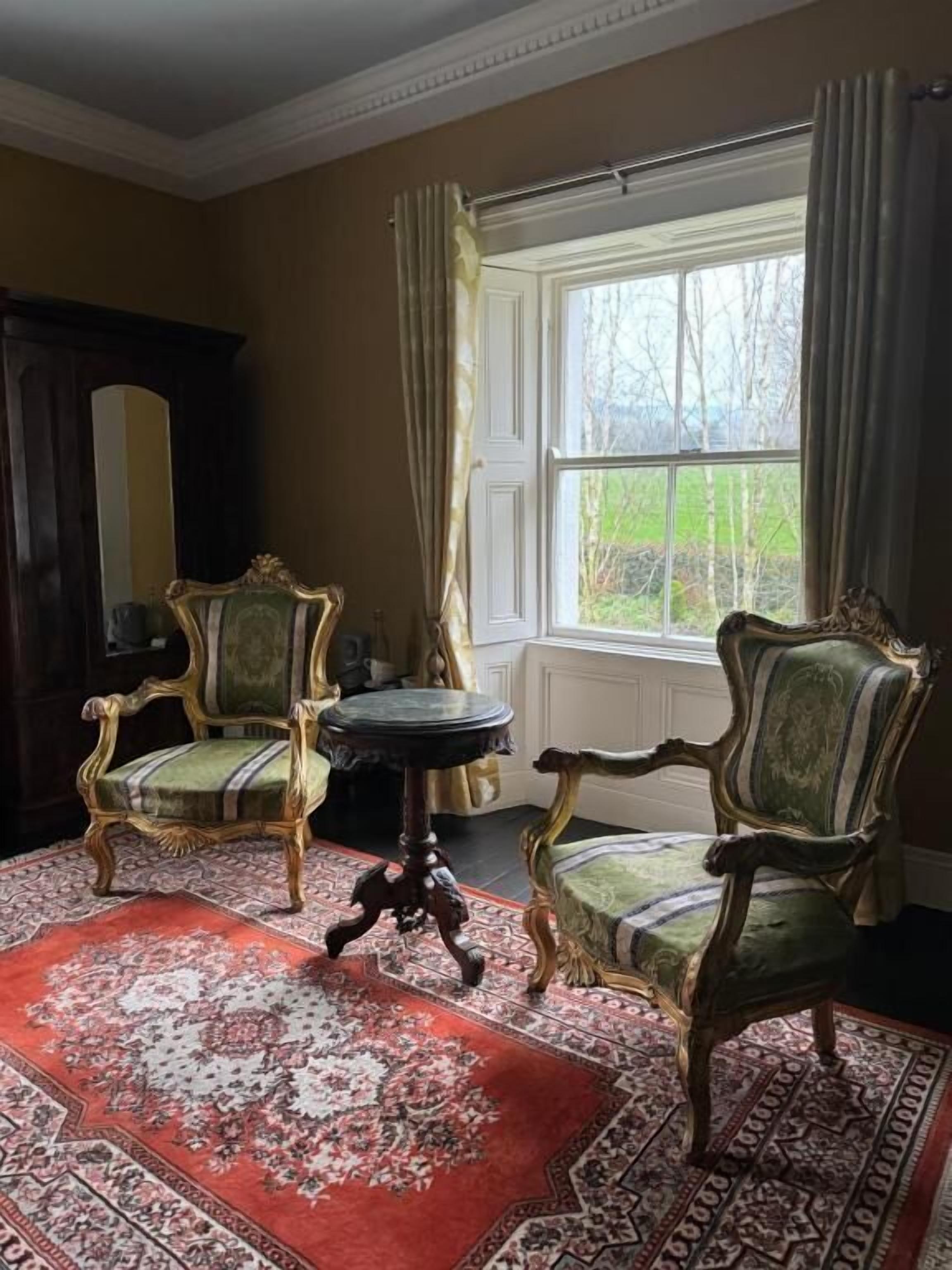 A room with two ornate chairs, a small round table, and a large window with curtains.
