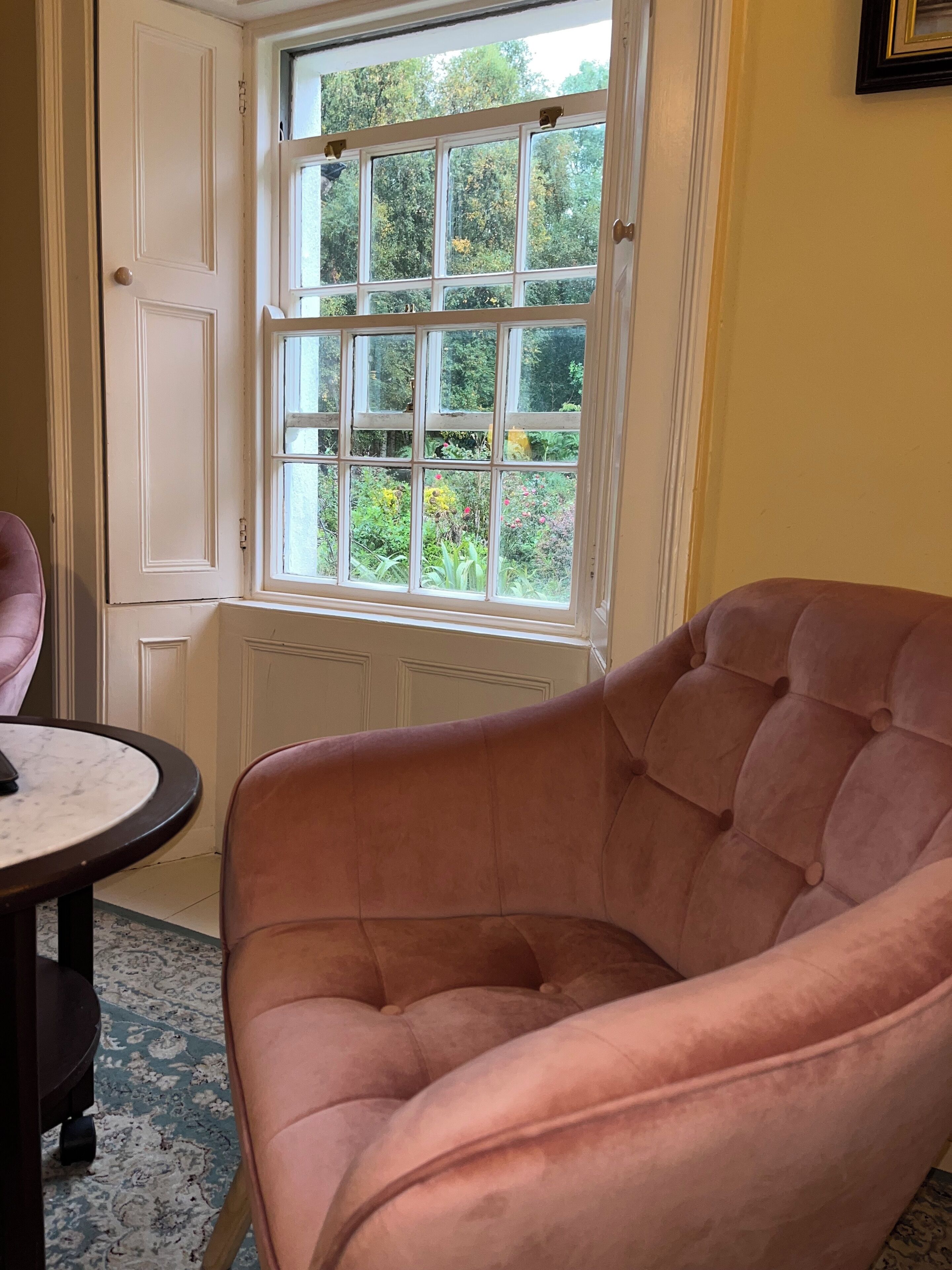 Una habitación con un gran ventanal, un sillón tapizado en rosa, una mesita redonda y una alfombra estampada.