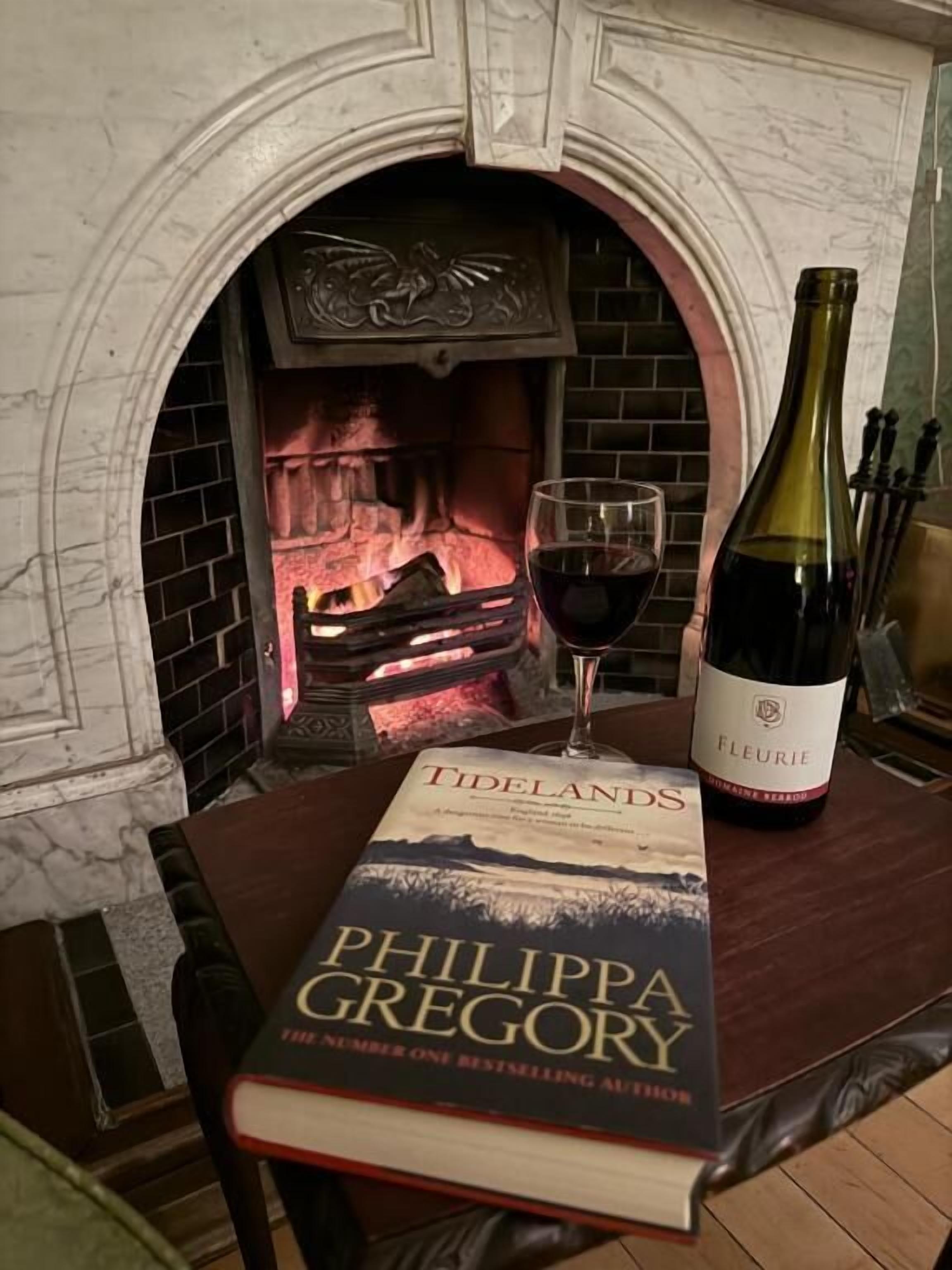 Een boek getiteld 'Tidelands' van Philippa Gregory, een fles wijn en een glas rode wijn op een tafel voor een open haard.
