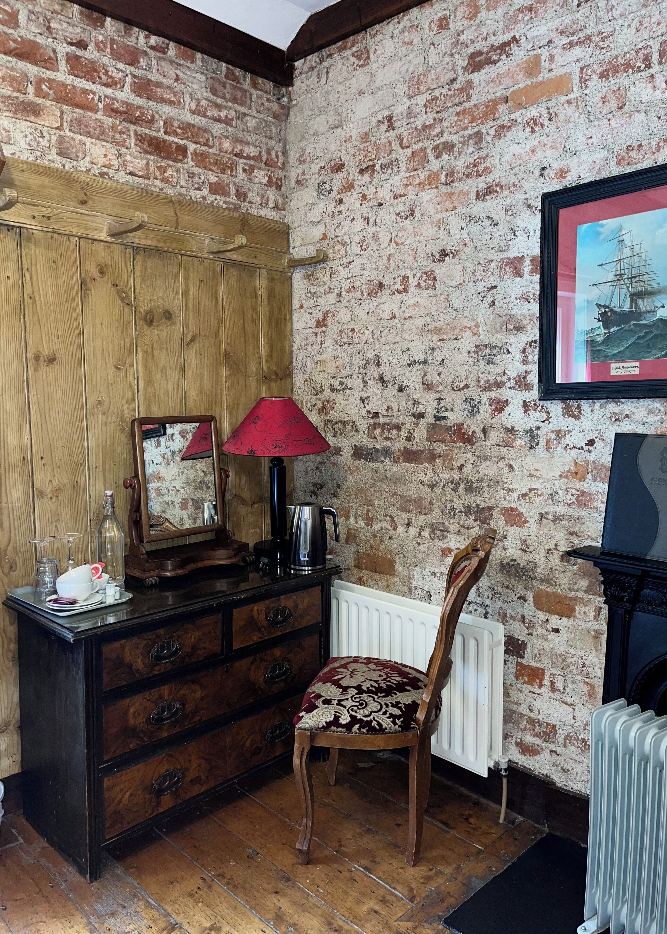 A room with a wooden dresser, a red lamp, a chair, a mirror, and a framed picture of a ship on the wall.