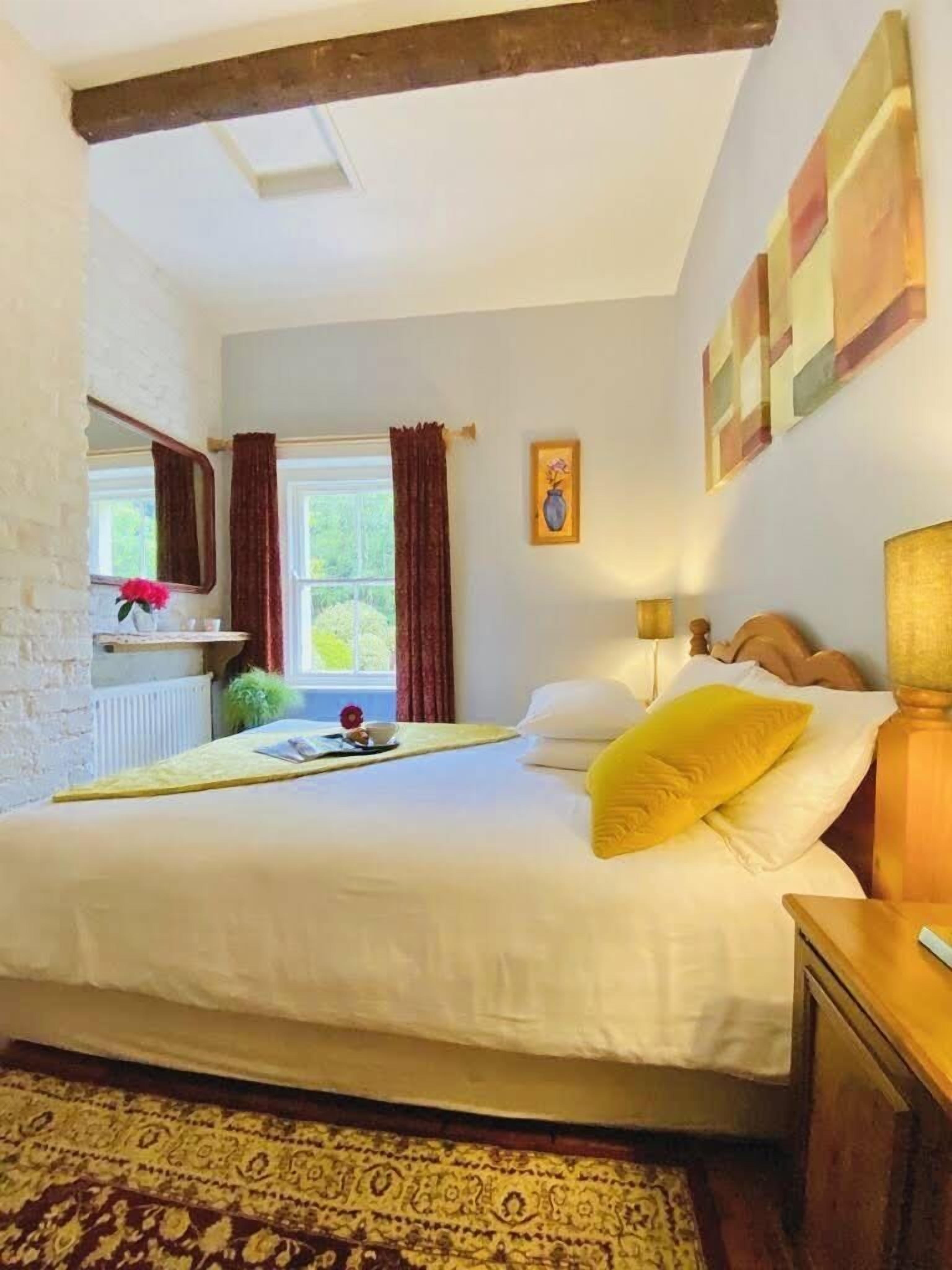 A bedroom with a large bed, a wooden headboard, a bedside table, a nightstand, a window with curtains, a radiator, and a wall-mounted painting.