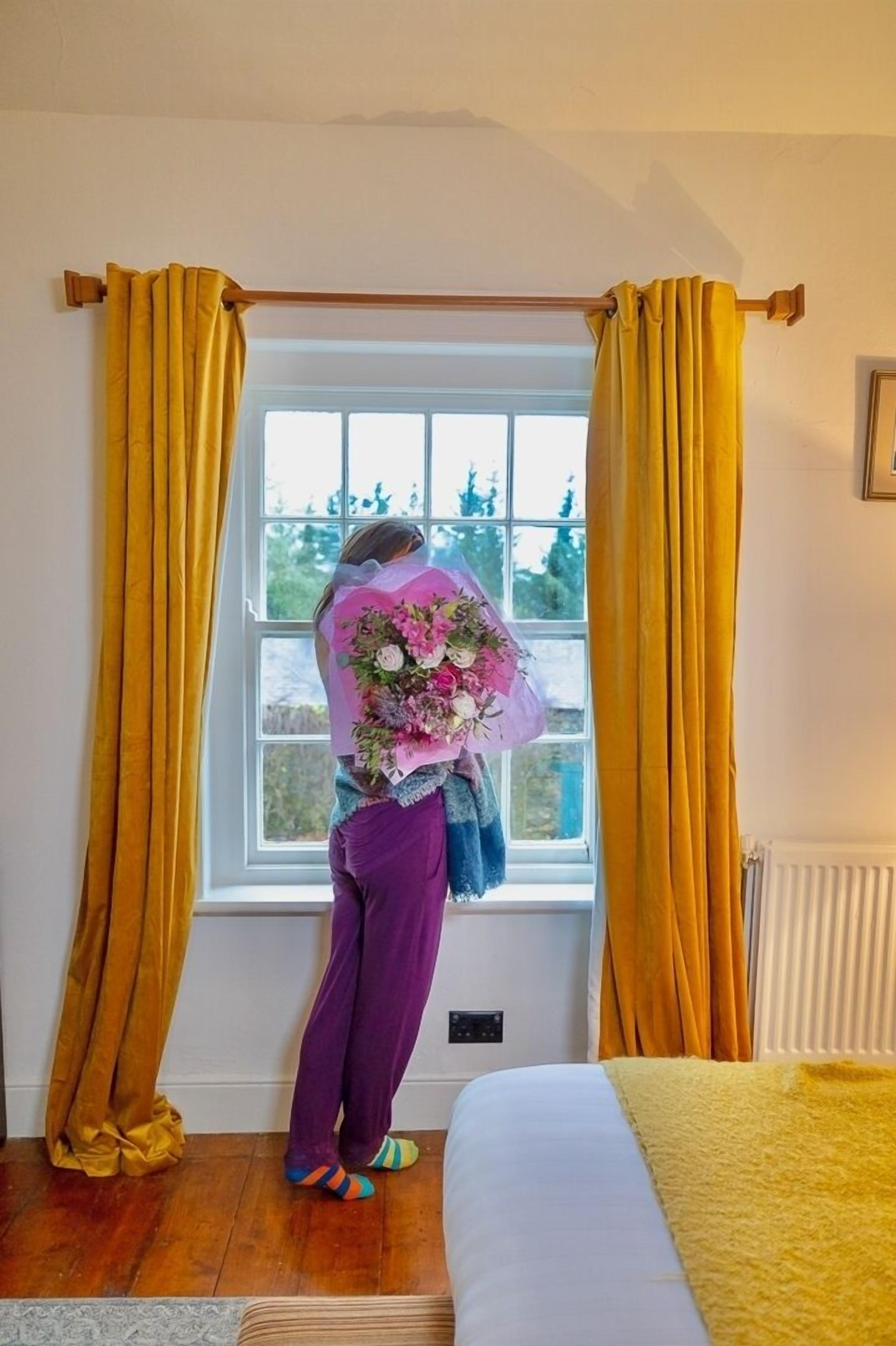 Una habitación con una cama, una ventana con cortinas amarillas y una persona junto a la ventana sosteniendo un ramo de flores.