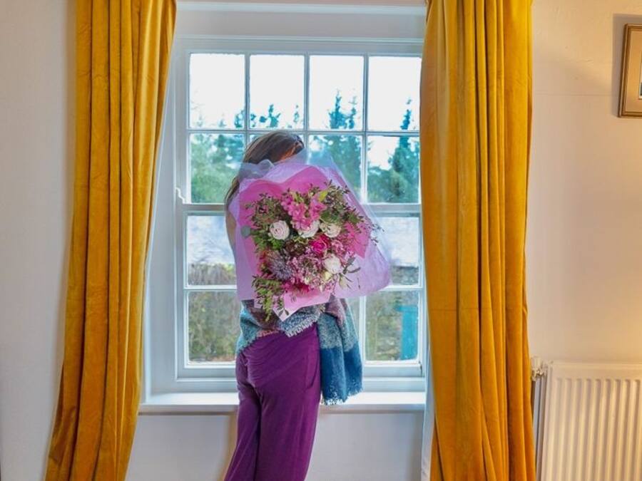 Una habitación con una cama, una ventana con cortinas amarillas y una persona junto a la ventana sosteniendo un ramo de flores.