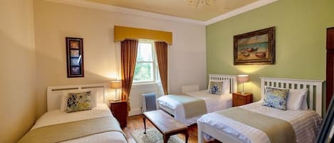 A hotel room with two single beds, a bench, a radiator, a window with curtains, a painting on the wall, and a chandelier.