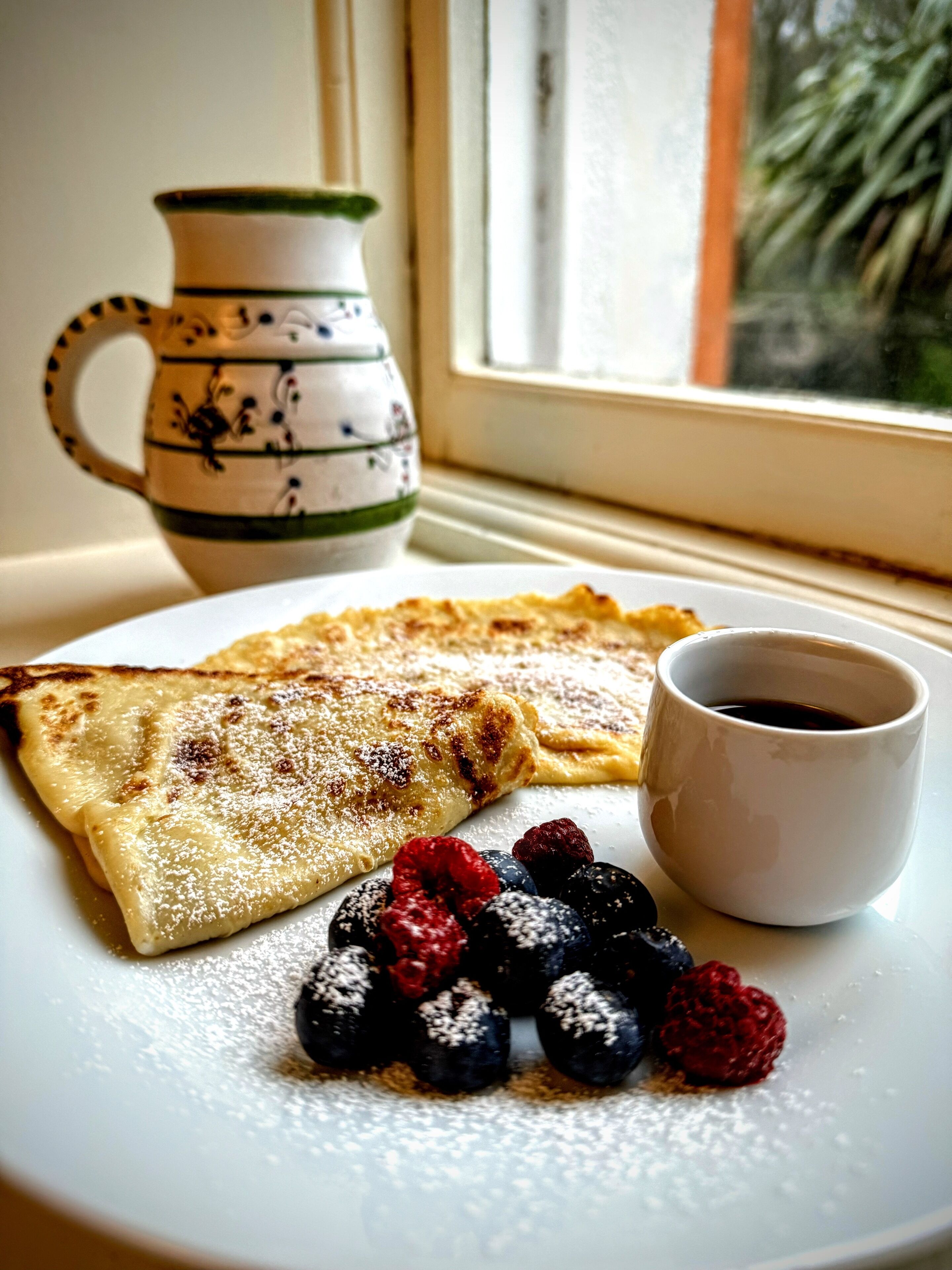 A plate with a crepe, berries, and a cup of coffee on a table by a window.