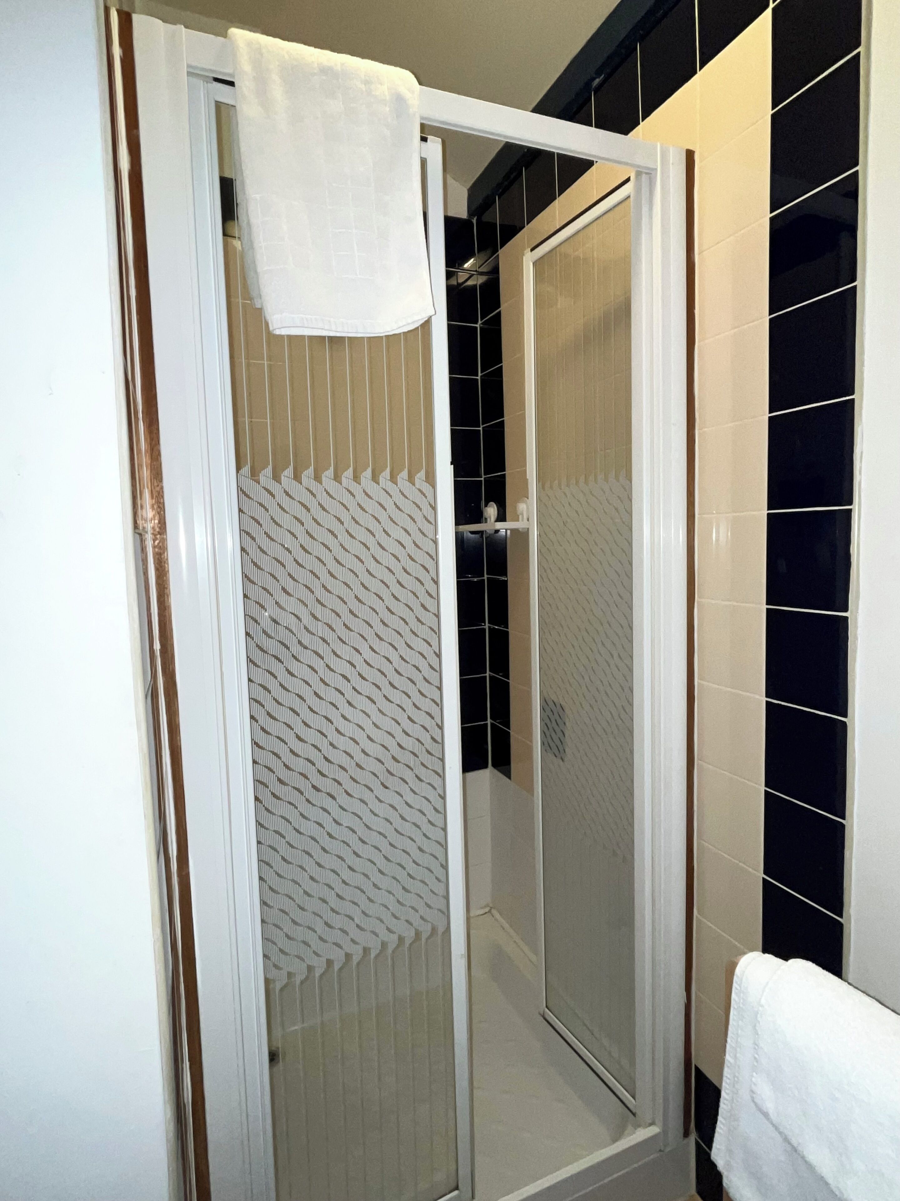 A shower enclosure with a glass door, a white towel hanging on the door, and a patterned shower curtain.