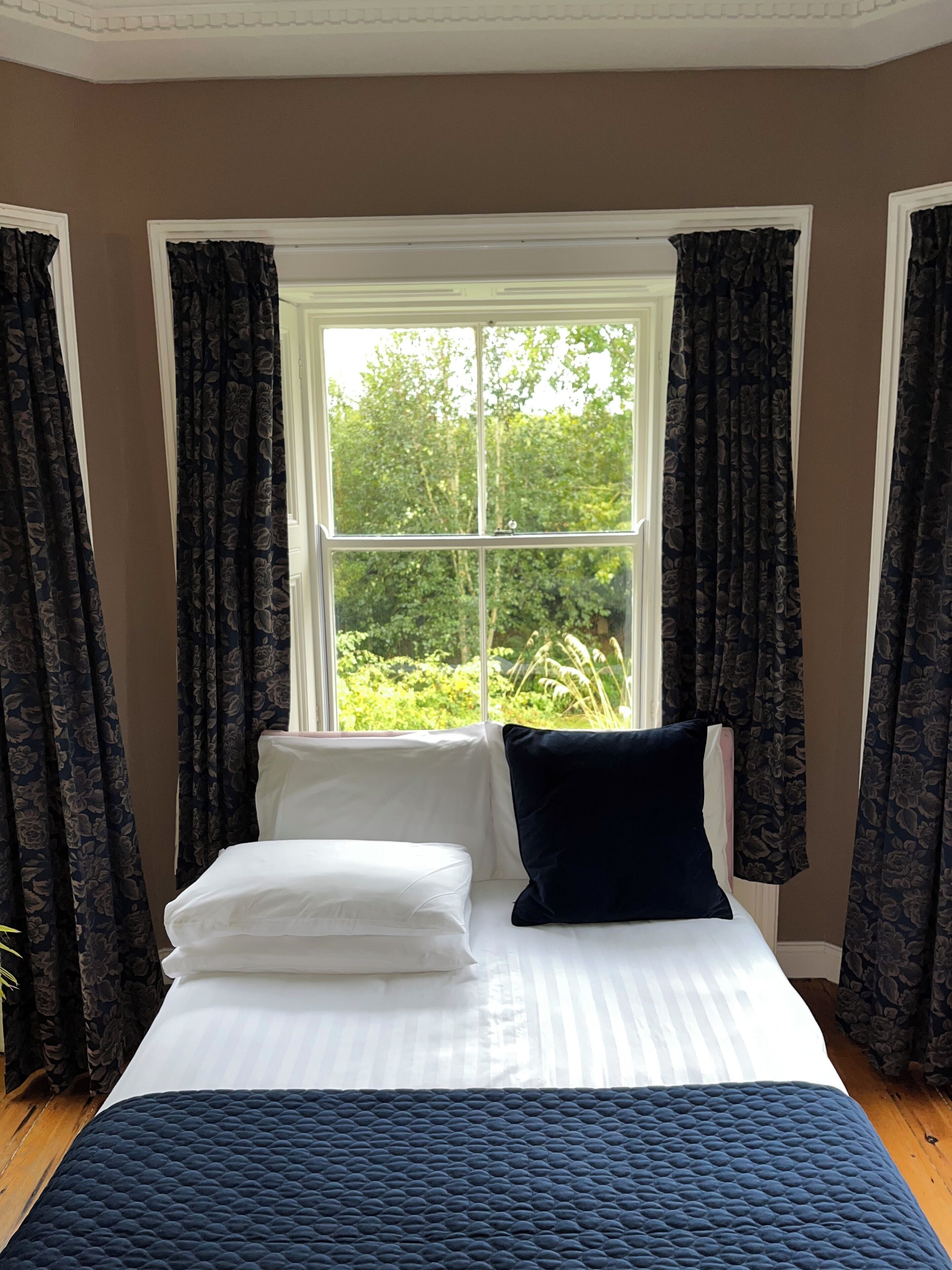 Un dormitorio con cama, ropa de cama blanca, colcha acolchada azul oscuro y una ventana con cortinas.