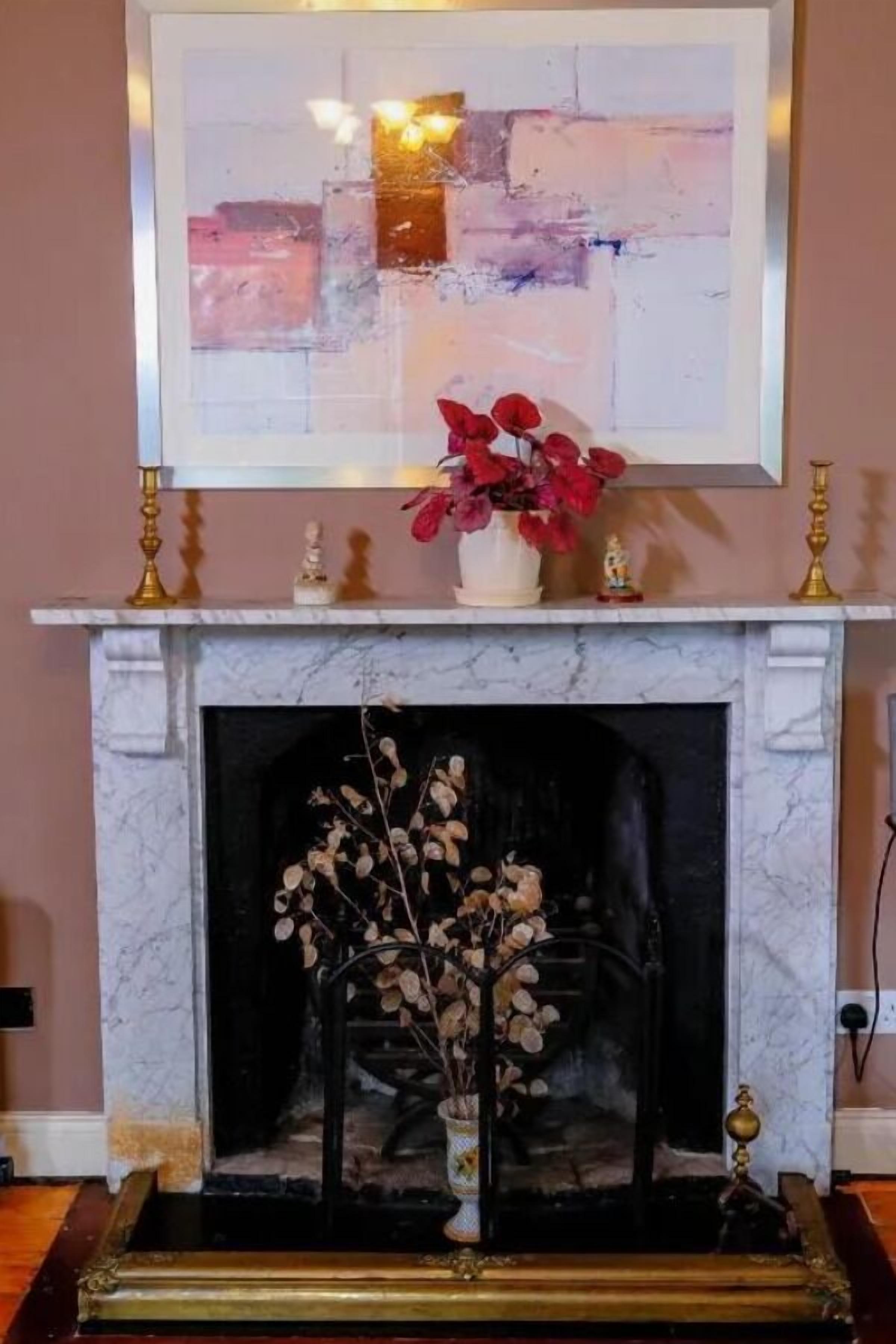 A fireplace with a white mantel, a framed abstract painting above it, and a vase with red flowers on the mantel.