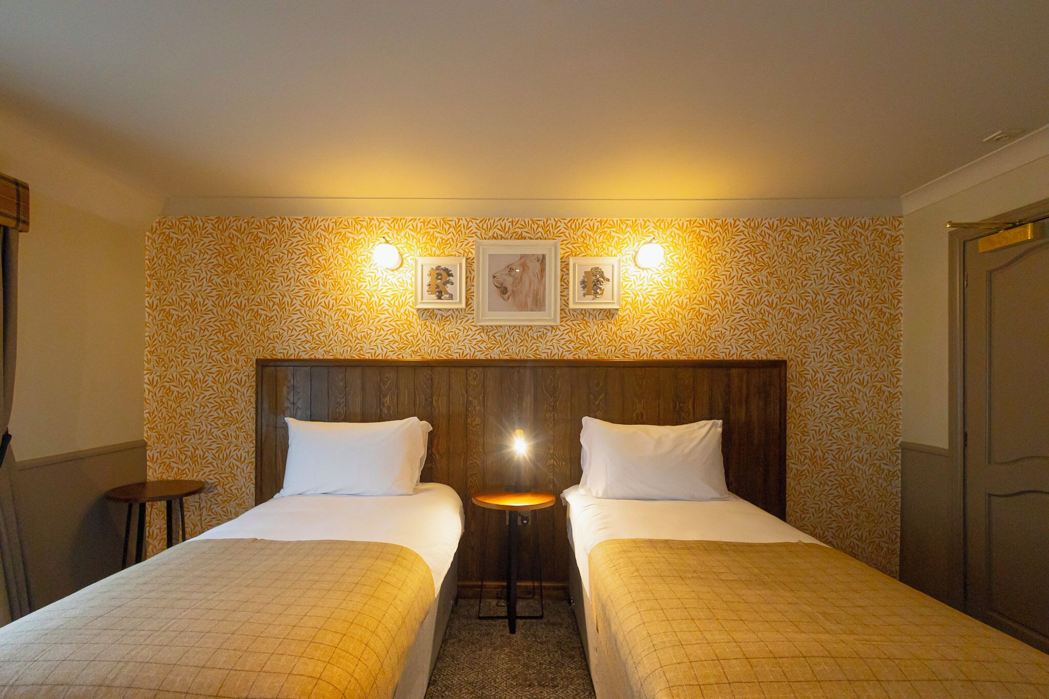 A hotel room with two beds, a small table, and a wall with patterned wallpaper and framed pictures.